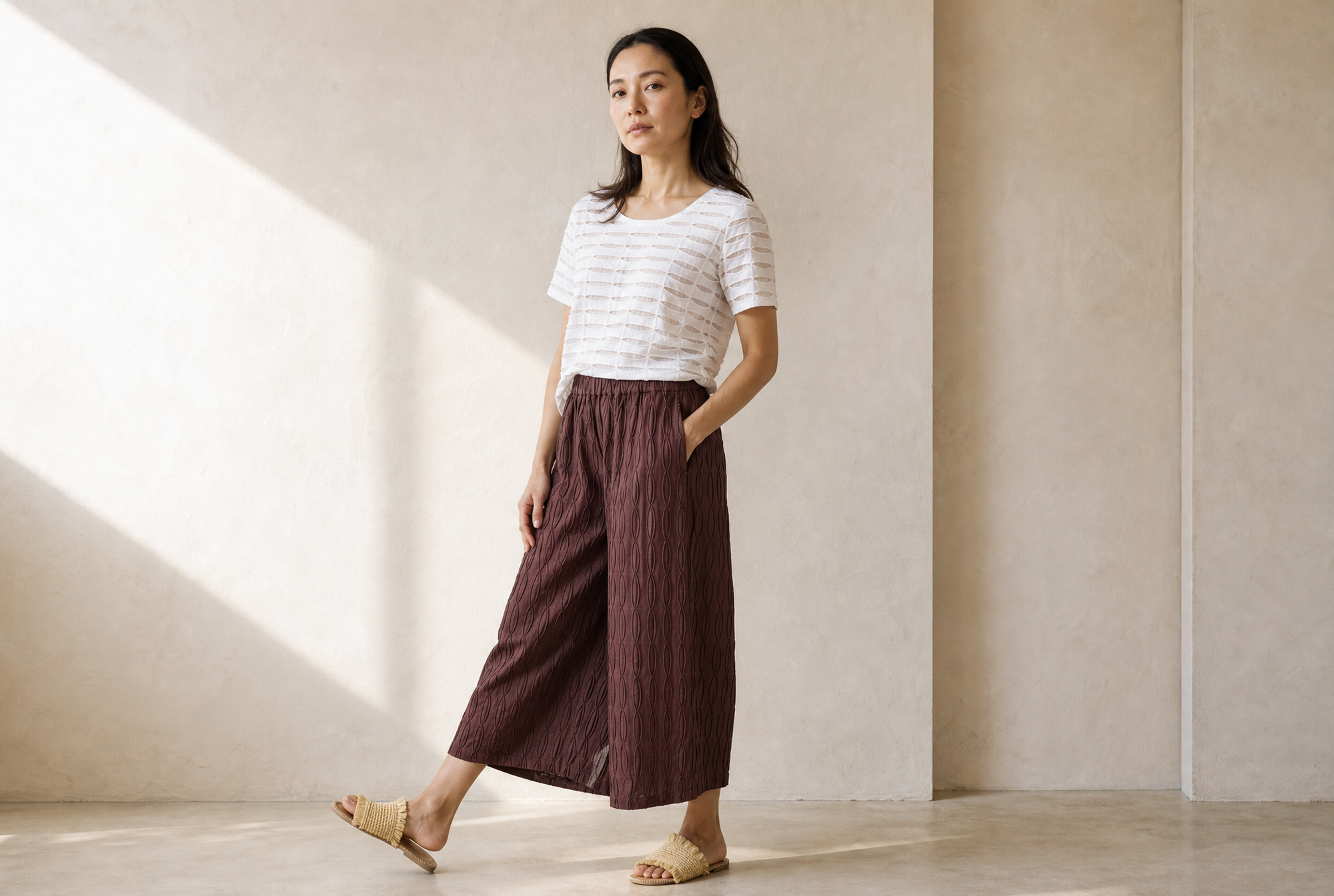 Woman wearing a white striped shirt and maroon pants standing in a minimalistic room with sunlight filtering through.